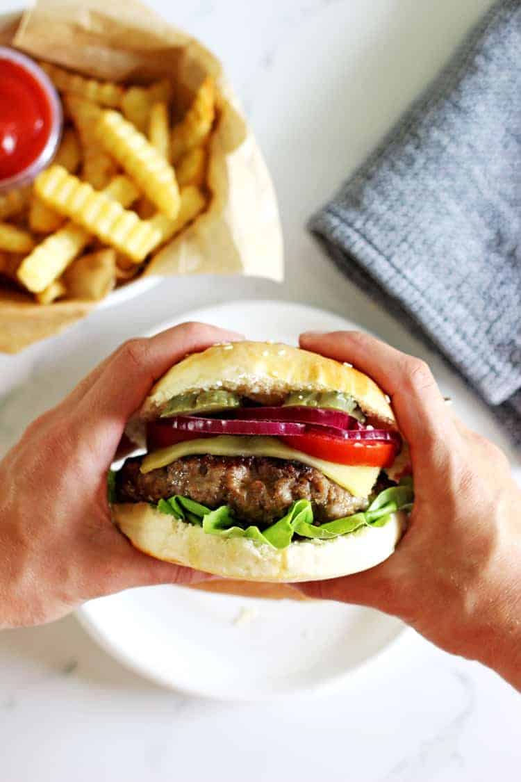 Hands holding huge burger with fries in the background